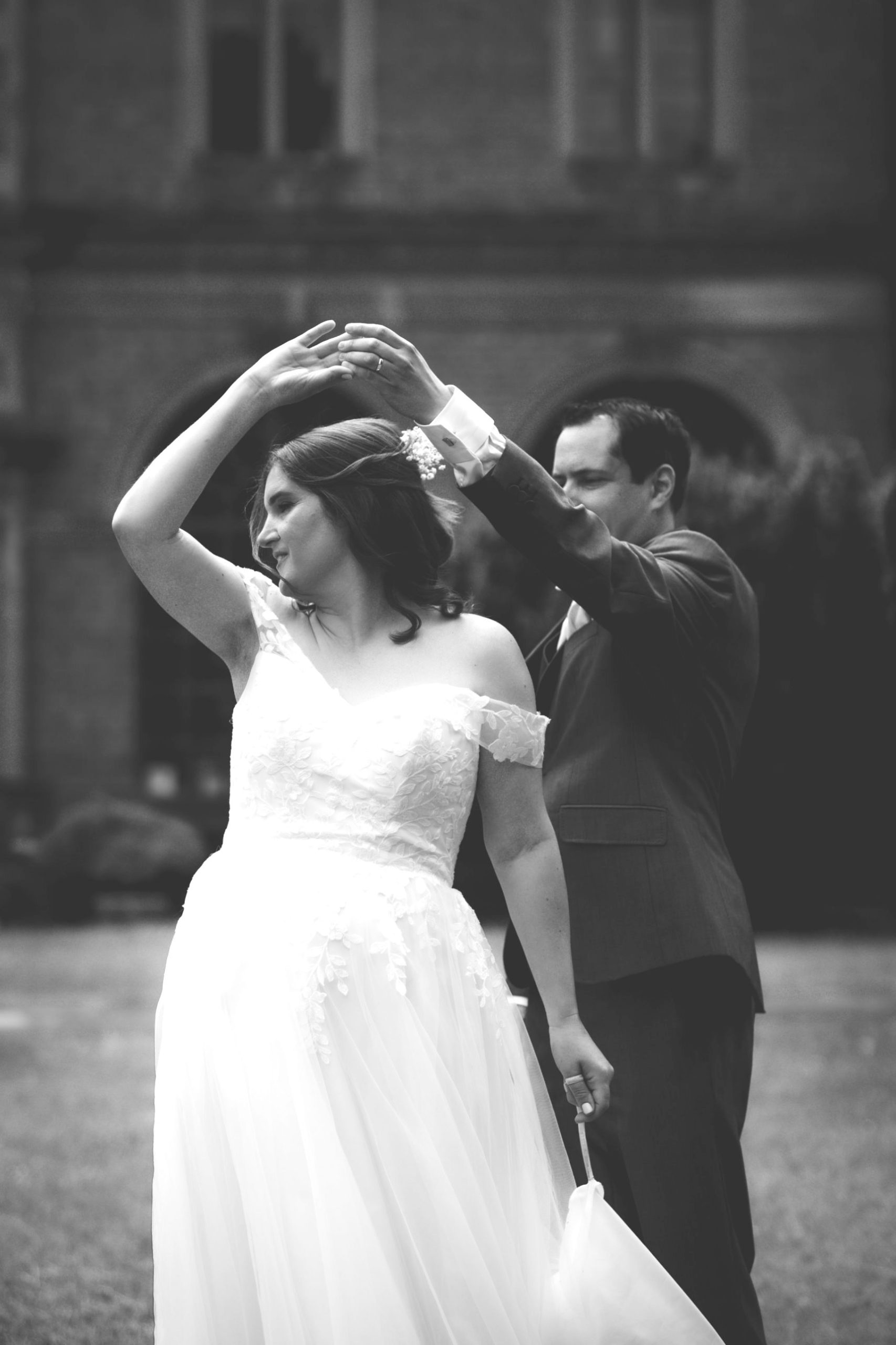 A bride twirls under her groom’s arm in front of Shaw House, in black and white